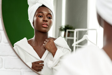 Woman in white bathrobe looking in mirror while adjusting towel on head in modern bathroom