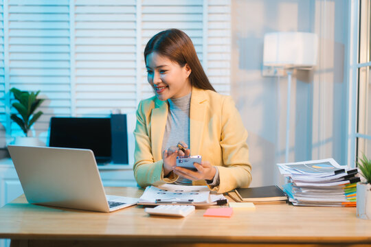 Asian woman works overtime at her office desk late at night, surrounded by paperwork, laptop, and smartphone, showing resilience and focus as she manages business tasks in a quiet
