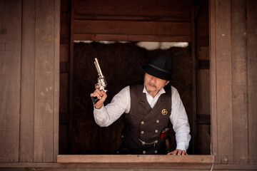 Portrait elder cowboy standing at a wooden window frame, aiming a silver revolver upward in a...