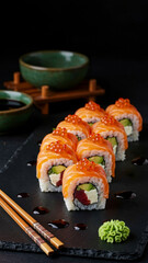 A row of sushi rolls with salmon, cream cheese, avocado, and tuna, topped with roe, served on a black slate with soy sauce, wasabi, ginger.