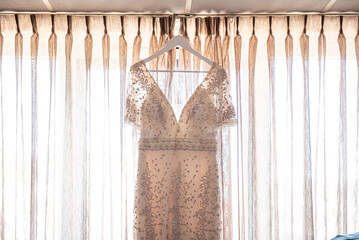 Beautiful champagne coloured weddingdress with little pajets and stones hanging in front of a window with transparent curtains insidea hotel room ready to be worn by the bride on the wedding day.