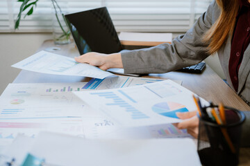 Young Asian businesswoman is holding documents and thinking about new project while working on laptop computer in modern office with multiple computer screens showing graphs