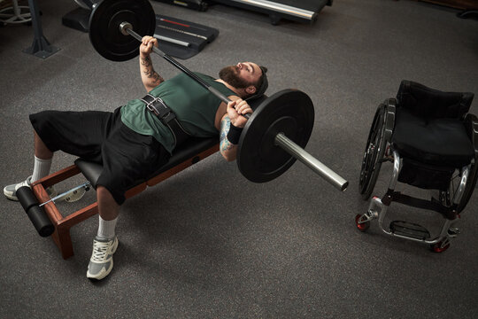 Man with tattooed arms lifting barbell weights while lying on exercise bench in gym. Wheelchair positioned next to the bench
