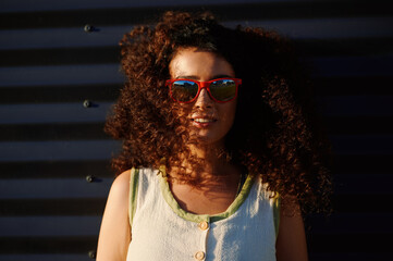 young woman with curly hair wearing fashionable red sunglasses standing against corrugated metal...