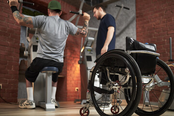Physical trainer assisting individual with exercise equipment in well-equipped gym. Visible...