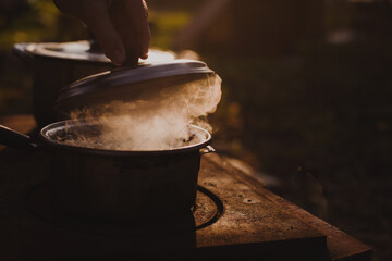 Lid of a steel pot being lifted above boiling water while cooking outdoors