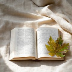 Fototapeta premium Open book on beige fabric with autumn leaves, soft sunlight, cozy and peaceful mood, perfect for fall reading inspiration