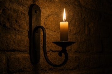 Single lit wax candle on rustic wrought iron wall sconce illuminating textured stone wall in a warm, dark interior