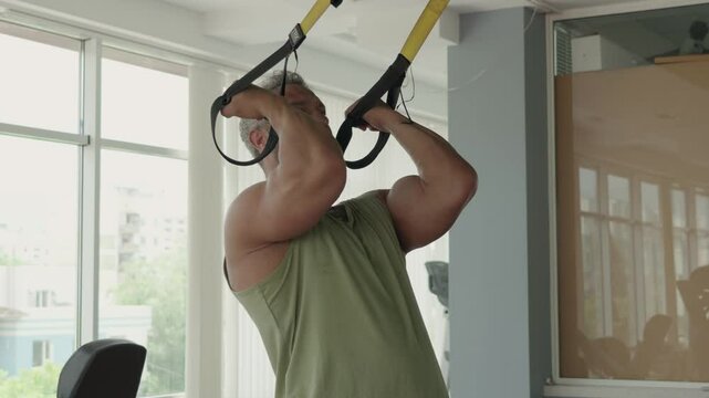 Man working out with suspension straps in a modern gym, focusing on upper body strength and fitness. Determined expression and muscular build.