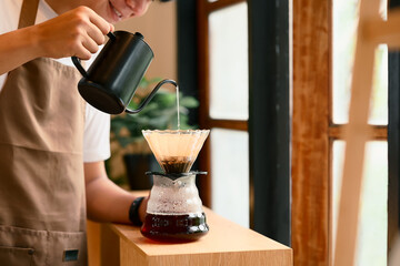 Young man in apron brewing coffee using the pour-over method in warm kitchen