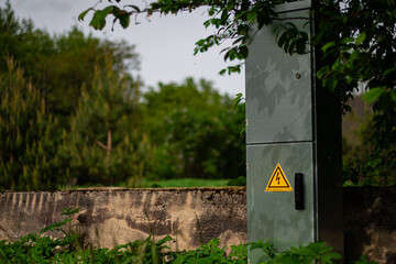 Metal electrical cabinet with a high voltage warning sign mounted beside a concrete wall, surrounded by overgrown greenery in a rural area.
