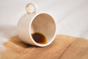 White ceramic cup resting on side with small coffee puddle on light fabric. Simple composition emphasizes spill, creating contrast between dark coffee and soft beige background
