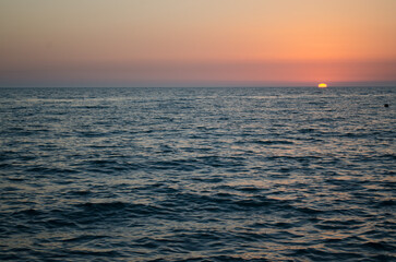 Obraz premium Calm ocean scene under warm, gradient sunset sky. Sun partially dips below horizon, casting orange glow. Peaceful atmosphere with gentle waves and expansive view