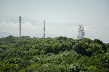 Obraz premium Ferris wheel stands tall among dense trees, with telecommunication towers nearby. Daylight highlights green foliage, while mountains fade in distant mist. Calm atmosphere captured from elevated angle