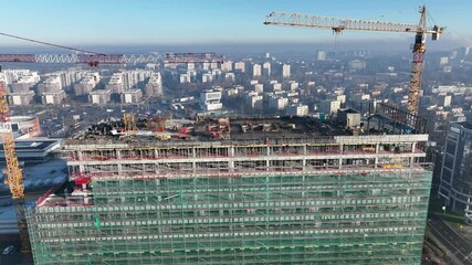 Aerial drone view of a modern high rise building under construction. Construction site with cranes working on a tall building with foundations and framework, workers, and machinery. Katowice, Poland.