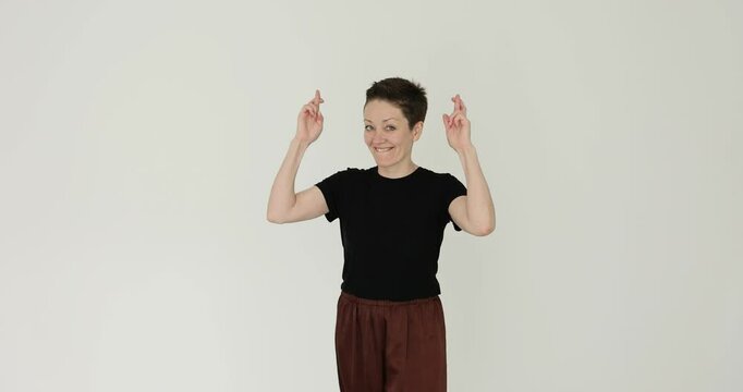 Young woman with short hair wearing t-shirt and pants crosses fingers and makes wish with hopeful expression. Anticipation and positive outlook
