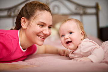 Young woman with brown hair and smiling baby girl on pink bed. Light-hearted interaction, creating joyful atmosphere. Warm lighting enhances bond