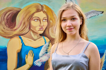 Young blonde female standing confidently before colorful mural. Artwork depicts woman, bird, blue...