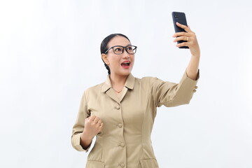 Excited young Indonesian civil servant woman holding a cell phone with fist hand. Isolated on white