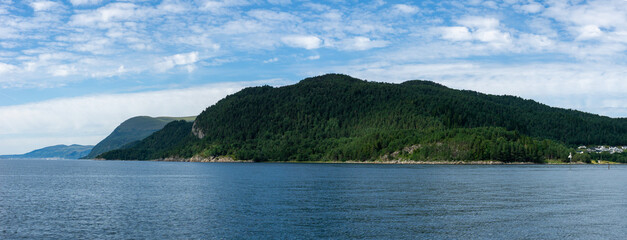 A lush green forest covers a coastal mountain along the Storfjord in Norway, near Solavagen, under a bright blue sky with scattered clouds on a calm, clear summer day