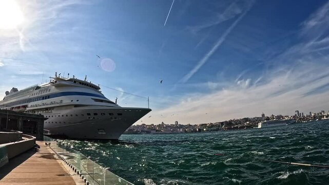 Wide-angle view of a large cruise ship docked in Istanbul's lively harbor. Clear blue skies, sun illuminating the scene, and waves gently lapping against the boat. Urban skyline in the background