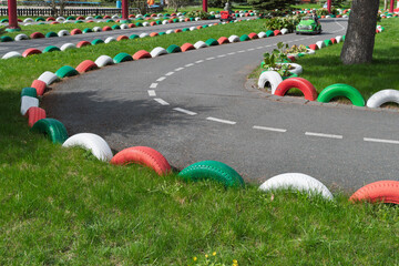 Ornamental racing track in park features winding asphalt road bordered by colorful tires on green grass, inviting playful movement.