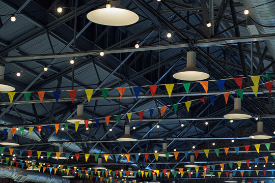 Bright festive flags and glowing string lights hang across modern industrial ceiling, setting lively mood for events in urban venue.