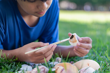 A young boy carefully cleans a seashell with a brush while lying on the grass, showcasing...