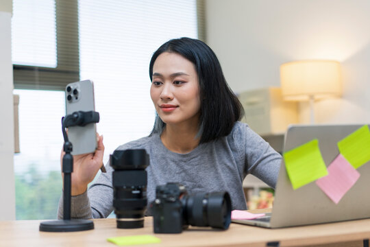 Young Asian woman engaged in content creation at home workspace with camera and smartphone
