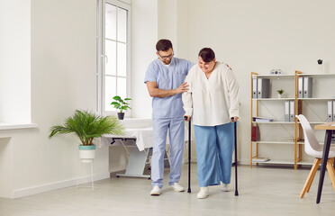 Fototapeta premium A young cheerful friendly nurse helping fat young patient to walk with her crutches in medical clinic. Male physiotherapist or health care worker helping woman in rehab.