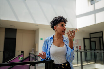 Young woman sending a voice message on her smartphone while standing in a modern, light filled...