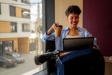 Smiling young creative woman with tattoos is comfortably working remotely, enjoying a coffee while...