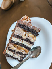 Layered chocolate cake slice on white plate with spoon. Rich dessert with creamy layers and nuts.