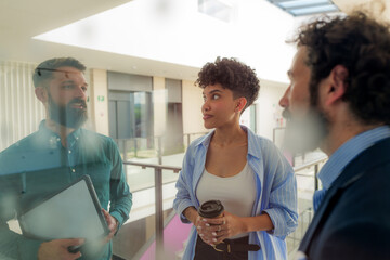 Three business colleagues are having a discussion in a modern office hallway, exchanging ideas and collaborating on a project, holding a tablet and a coffee