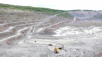 A large quarry is in full operation with heavy machinery moving earth and rocks. The rugged terrain highlights both the excavation process and the surrounding environment