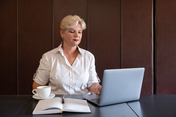 Mature businesswoman working on her laptop at a cafe, professional person focused on the screen of a laptop with a cup of coffee aside and an open planner in a calm and productive work environment