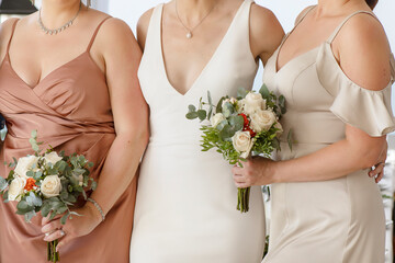 3 elegantly dressed women, likely a bride with a deep v neckline dressed and her bridesmaids in satin and light beige gowns, holding small bouquets of cream and peach roses with eucalyptus greenery