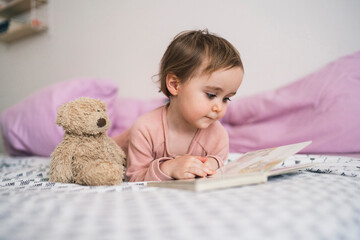 A young child lies on a bed with a teddy bear companion, engrossed in looking at a book. This quiet moment shows early childhood learning and relaxation.