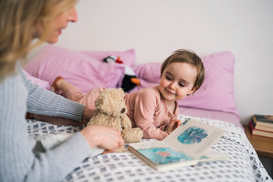 A mother reads a storybook to her happy toddler on the bed. They share a moment of connection and learning during quiet time at home.