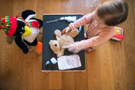 A young girl plays doctor, caring for her teddy bear patient. She uses toy medical tools and a diaper on a small table.