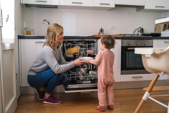 A mother and her young child are in the kitchen. The mother kneels by the open dishwasher and hands a plate to the child. They are doing chores together.