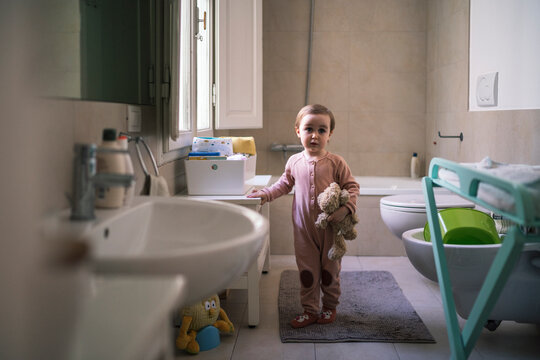 A young child stands in a bathroom holding a teddy bear. The toddler wears a pink jumpsuit and looks directly at the camera. The bathroom is tidy with a sink, toilet, and bathmat.
