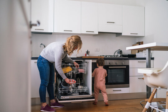 A mother is busy loading the dishwasher with dirty dishes in her modern kitchen. Her small child is standing close by, watching her. Everyday family routine.