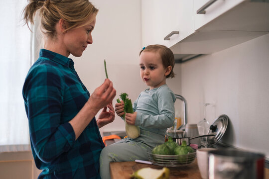 A mother and her young daughter are in the kitchen preparing vegetables. The child holds a fennel bulb while the mother shows her a green stalk. They share a moment while cooking.