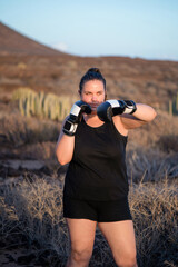 Confident woman wearing black sportswear and boxing gloves poses in a determined stance among towering cacti in an outdoors natural setting with a rugged landscape, sports practice showing resilience