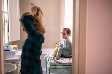 In the bathroom, a mother does her hair while her little daughter sits on a changing table, observing her. A moment of family life during the morning routine.