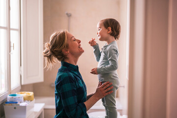 Mother and young child share a happy moment at home. The child stands while the mother looks up and smiles, holding the child's legs. They are enjoying time together.