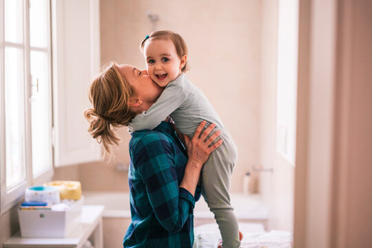 A mother holds her adorable toddler daughter and gives her a sweet kiss on the cheek. The happy child smiles and looks at the camera indoors.