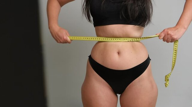 Woman tightly measuring waist with tape in front of mirror, body image pressure and weight loss concept.