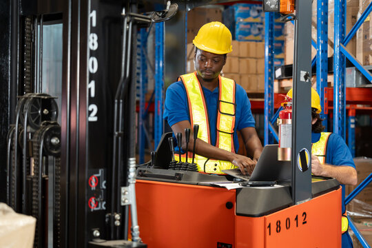 Black male warehouse worker in helmet and reflective vest operating forklift control panel while using laptop, with another male colleague partially visible behind, inside storage facility 
